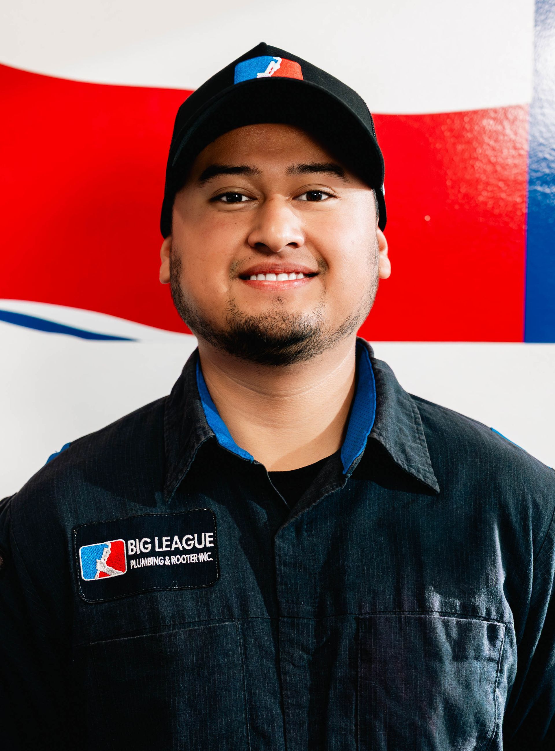 Person in a black cap and jacket standing before a red, white, and blue wall logo.