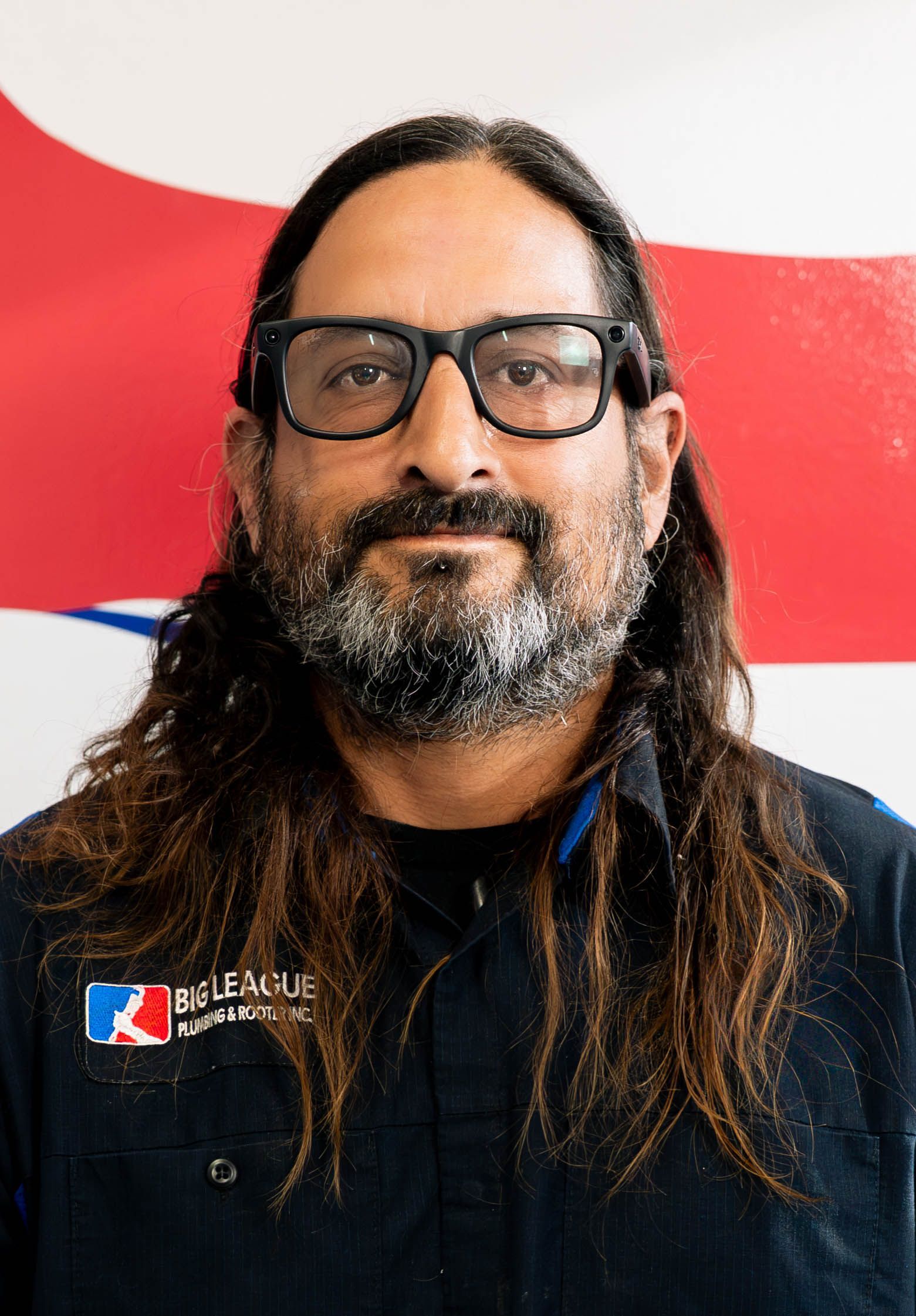 Portrait of a person with long hair, glasses, and a beard, wearing a black shirt against a red and white background