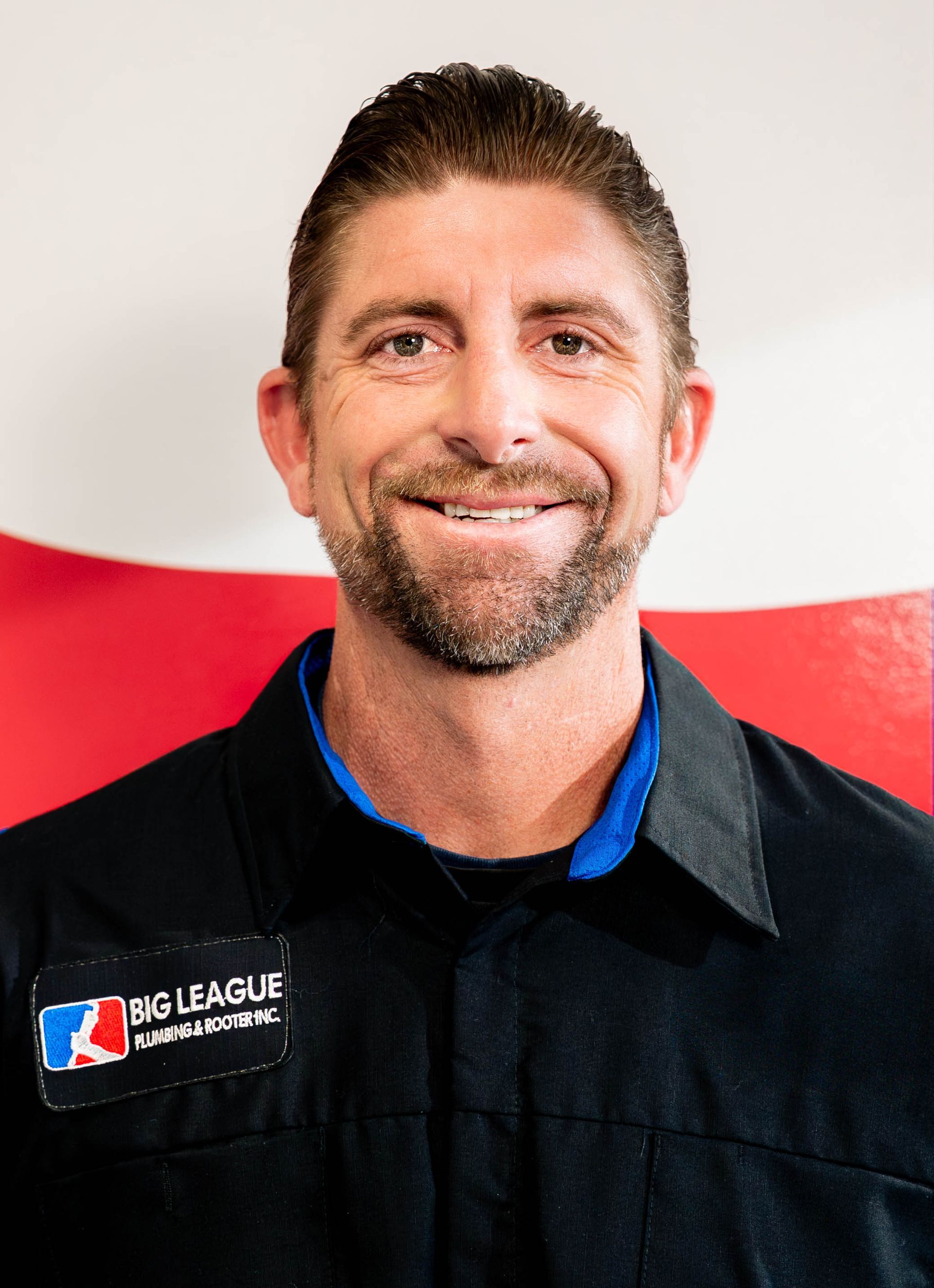 Smiling man in a black work shirt with a logo, standing before a white and red background.