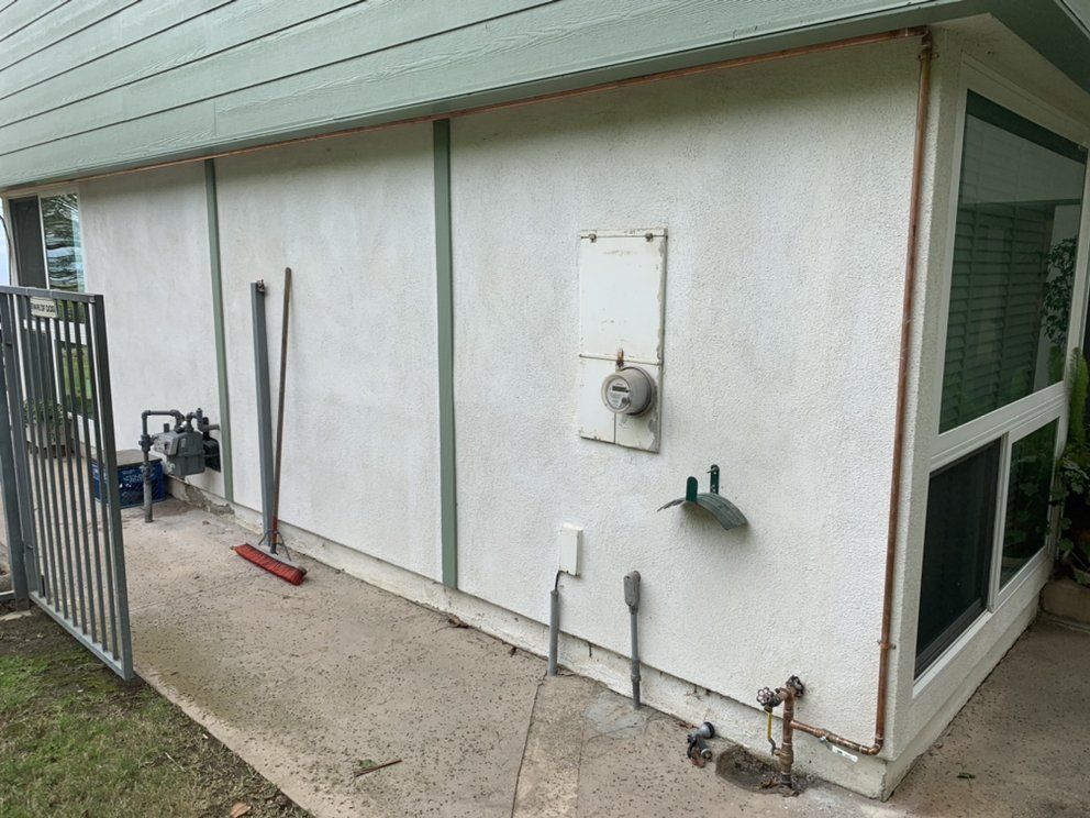 Exterior side of a white utility building with pipes, a meter, and a concrete walkway beside a fenced yard
