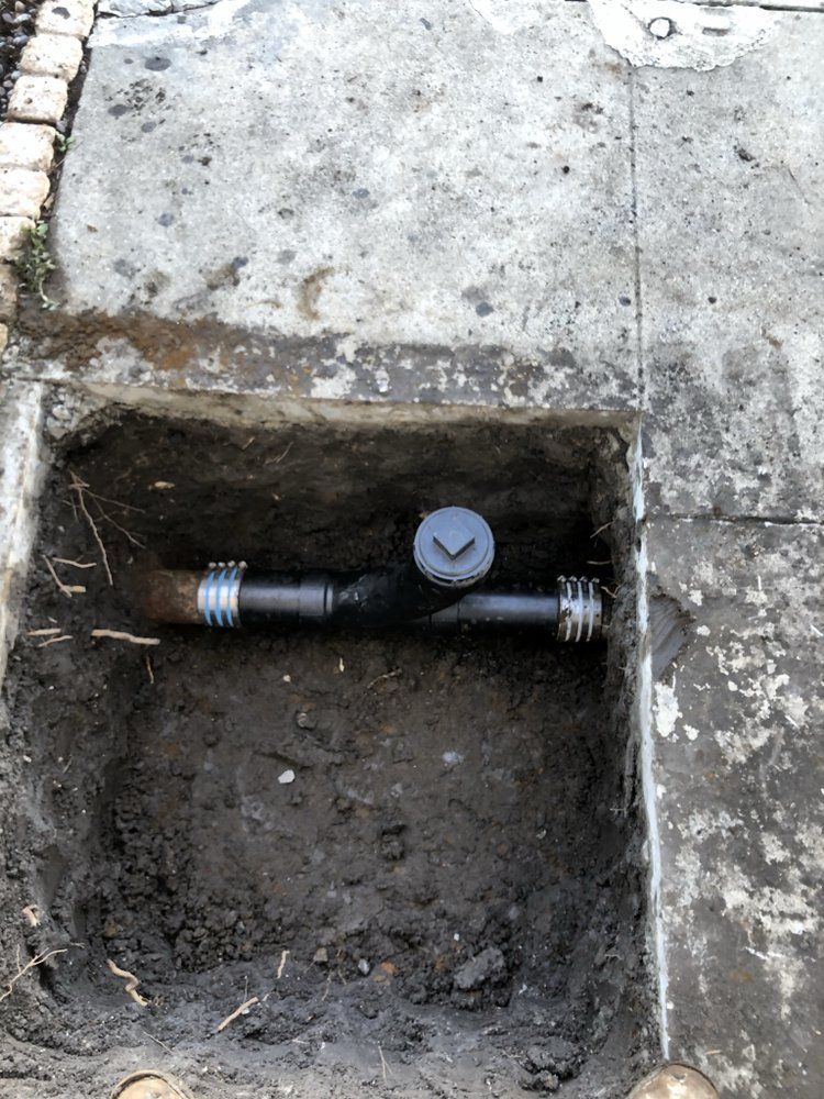 Open concrete utility box with a black pipe and capped fittings in a dirt-lined hole
