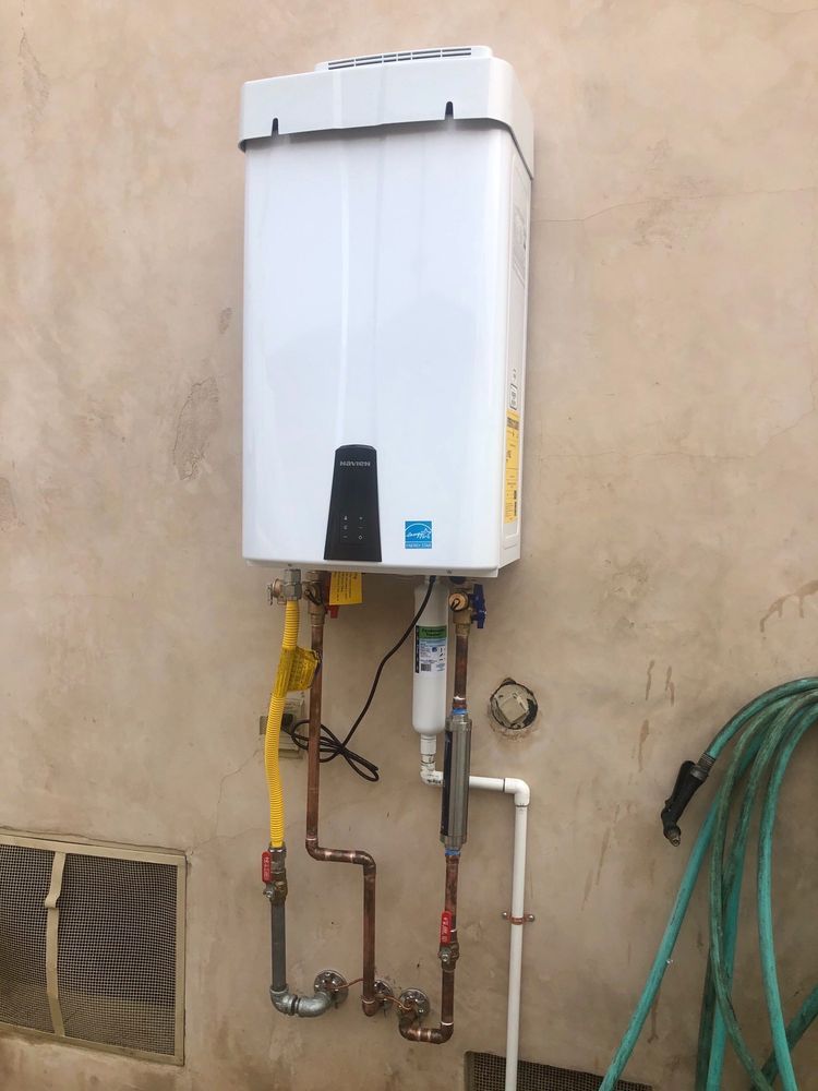 Wall-mounted white gas water heater with exposed pipes on an outdoor beige wall.