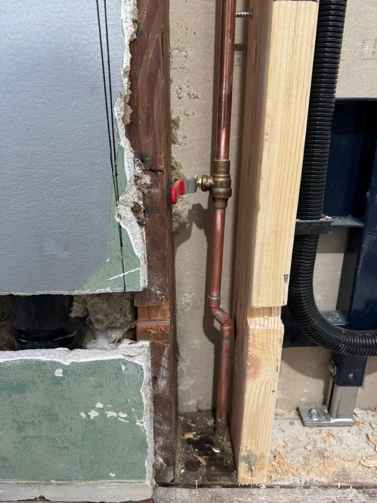 Wall opening exposing copper pipes and a valve beside insulation and wood framing.