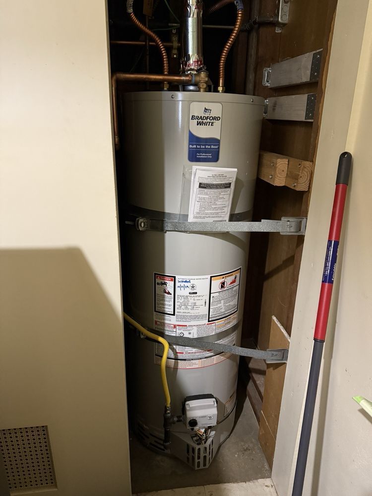 Tall water heater in a narrow utility closet with pipes, labels, and a gas line beside it