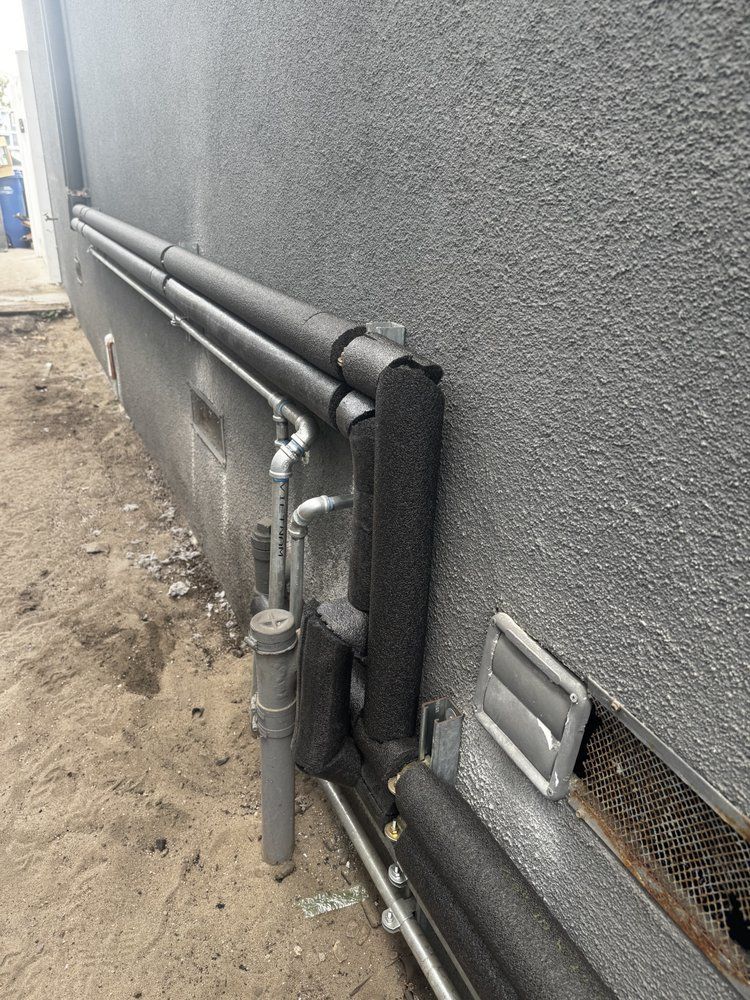 Gray exterior wall with exposed pipes and utility fixtures along a narrow side yard.