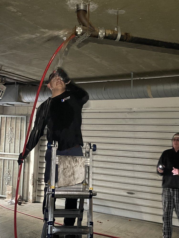 Person on a step ladder working overhead on exposed pipes in a garage, holding a red hose