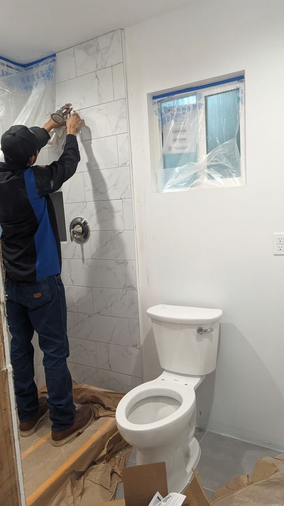 Worker installing wall tiles in a small bathroom with a toilet and window.