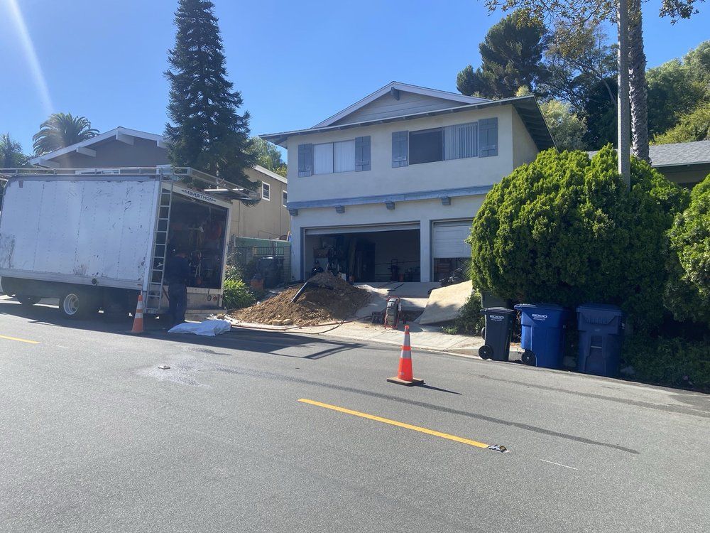 House with driveway excavation, a dump truck, and orange cone on a sunny suburban street