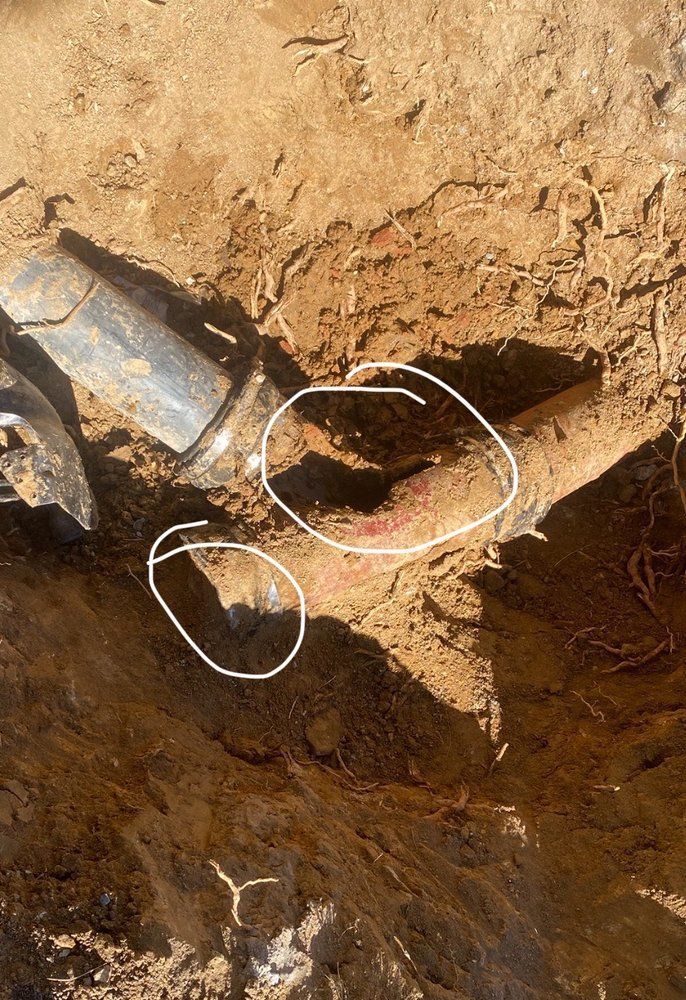 Excavator bucket digging a trench in dry soil, with a pipe or cable exposed and circled in white.