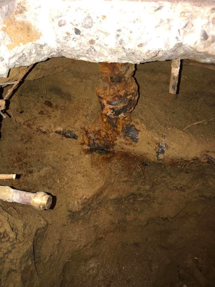 Rusty pipe and corroded metal beneath a concrete opening in a dirt crawl space
