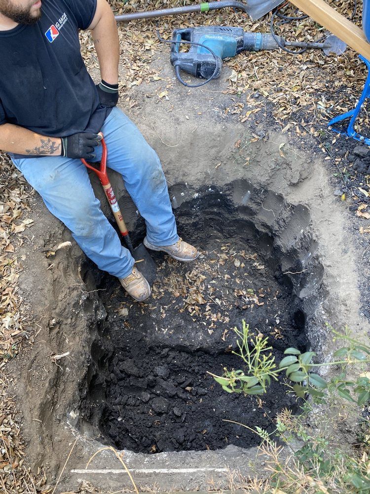 Worker standing in a deep dirt pit beside a blue machine, using a shovel or tool outdoors.