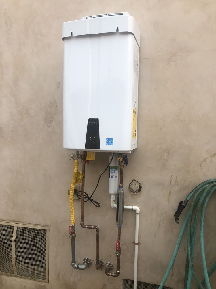 Wall-mounted white gas water heater with pipes and valves on an outdoor beige wall.