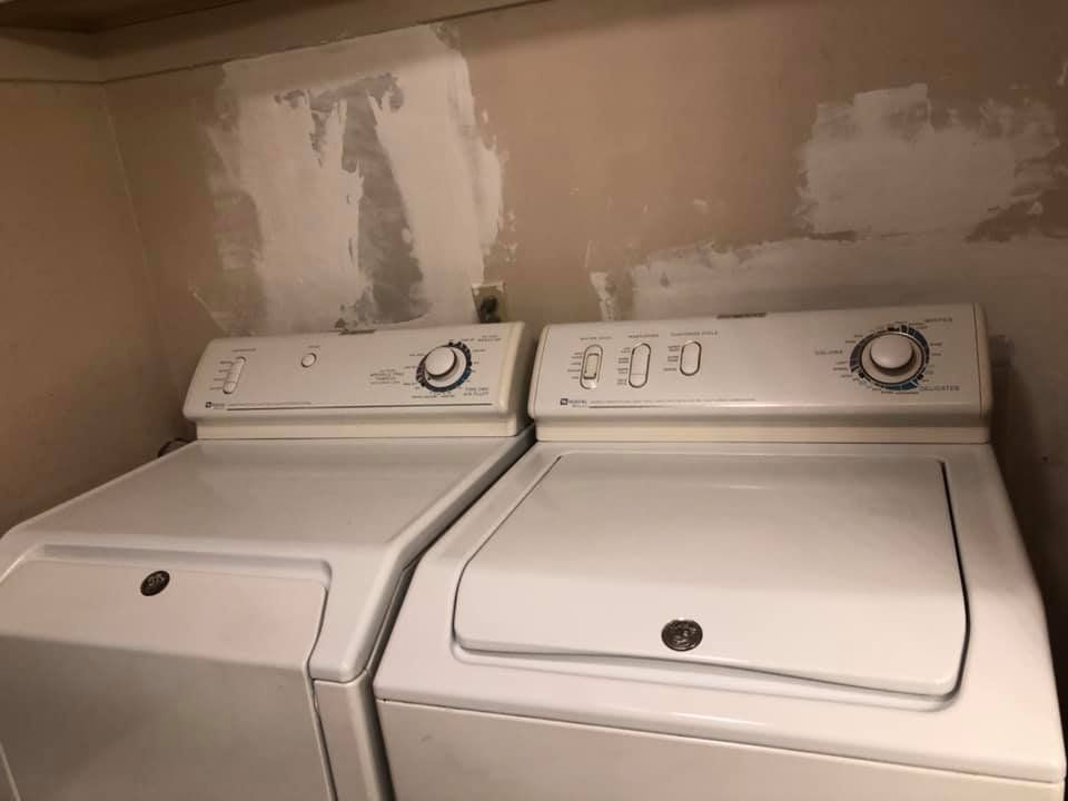 Two white top-load washing machines in a laundry room, with a beige wall behind them.