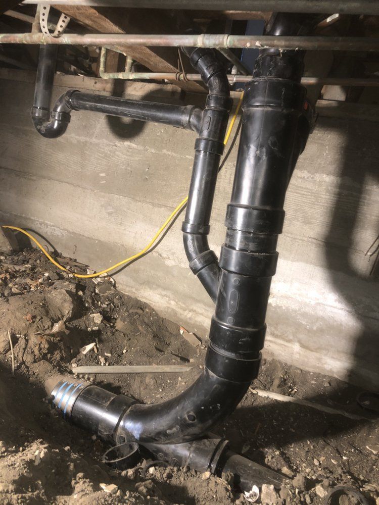 Black PVC drain pipes under a floor joist in a crawl space, with dirt and a yellow cable nearby