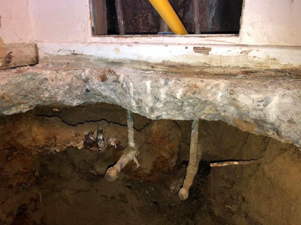 Broken floor exposing pipes and a hole beneath a wall opening