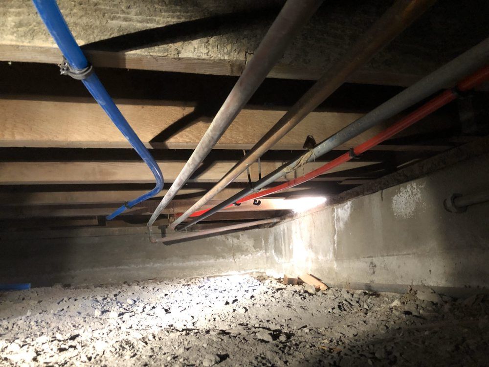 Crawlspace with exposed pipes and beams over a dirt floor, lit by a small light.