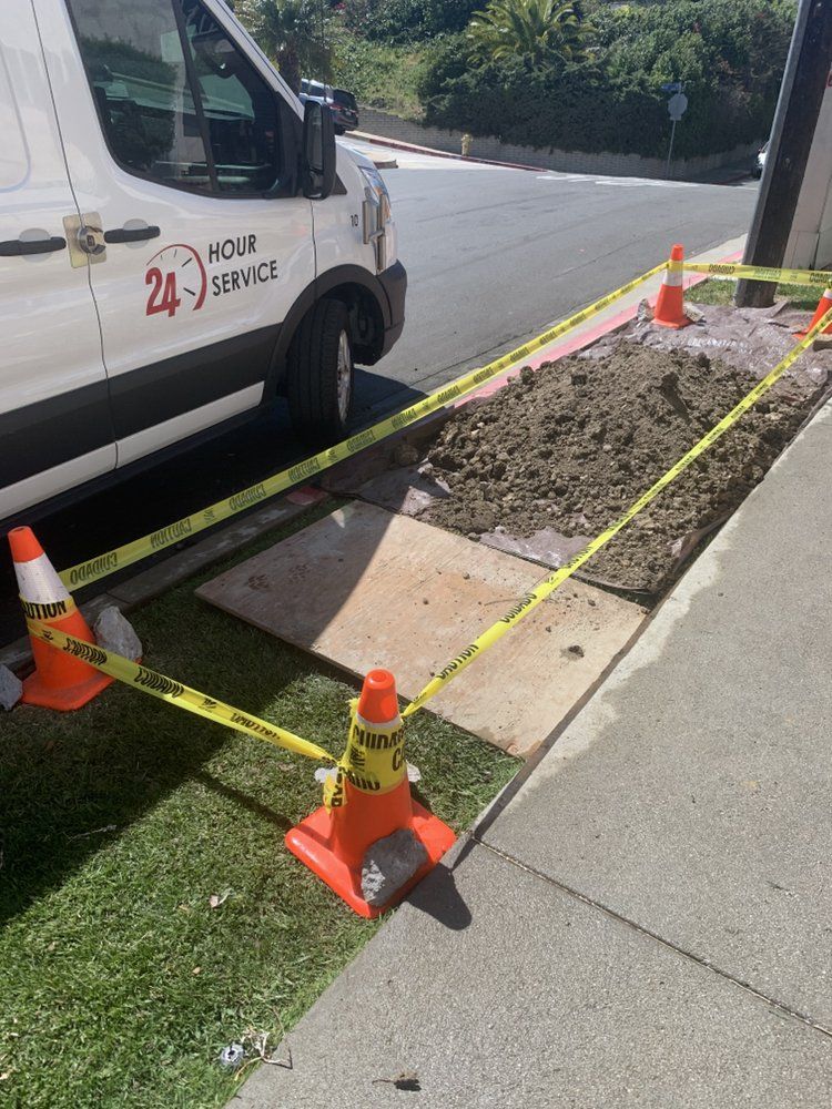 Curbside sidewalk excavation beside a white van, marked off with yellow caution tape and orange cones.