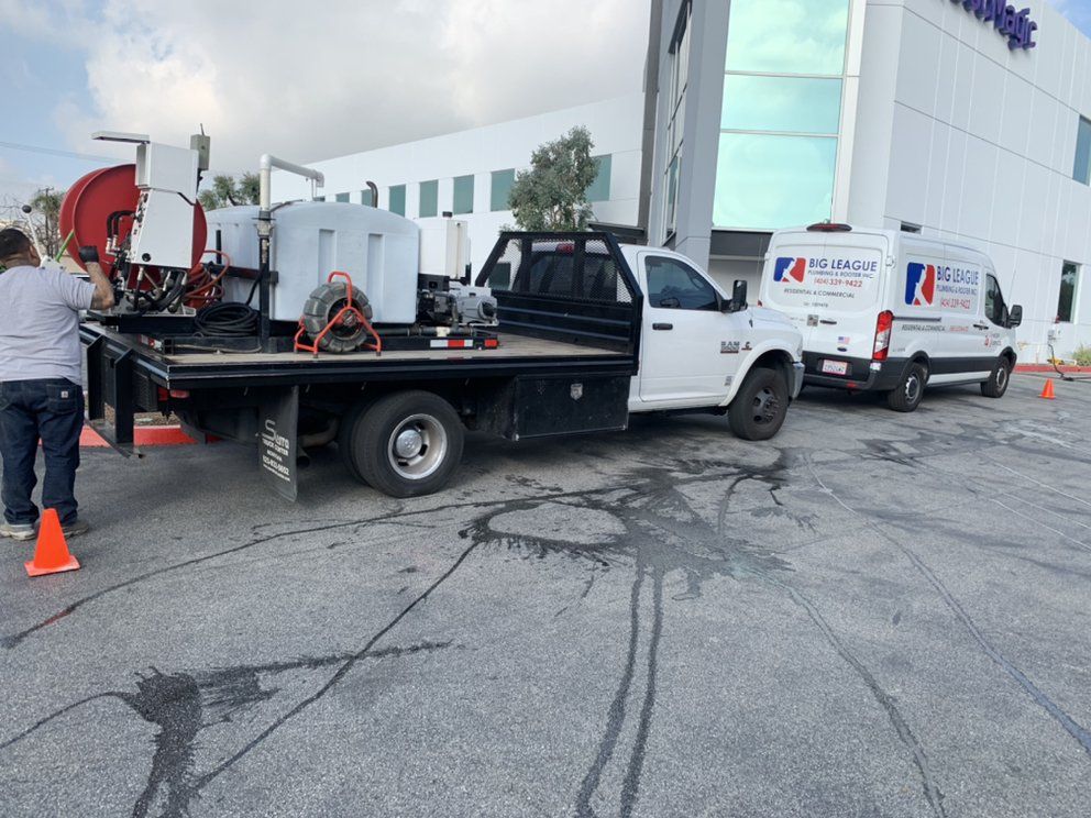 Utility workers with truck-mounted equipment beside a white building, with traffic cones on cracked pavement.