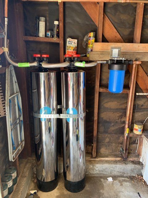Two chrome water softener tanks with a blue filter canister in an unfinished utility room