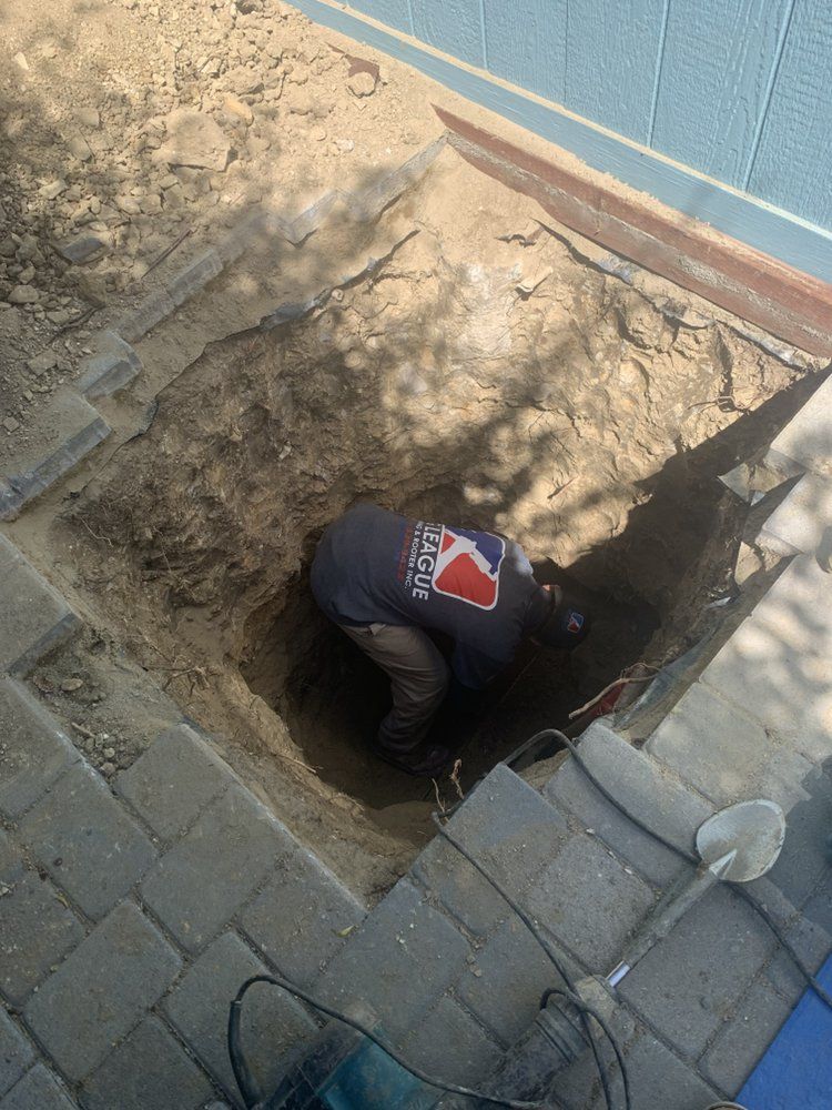 Worker in a trench beside a blue wall, digging near paved stones with a shovel.