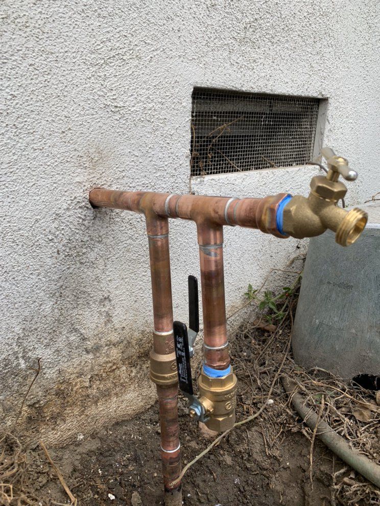 Outdoor copper water pipes and valves beside a stucco wall, with a window vent above.
