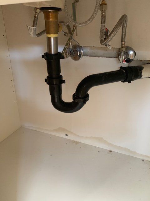 Under-sink plumbing with black drain pipes and metal water lines inside a cabinet.