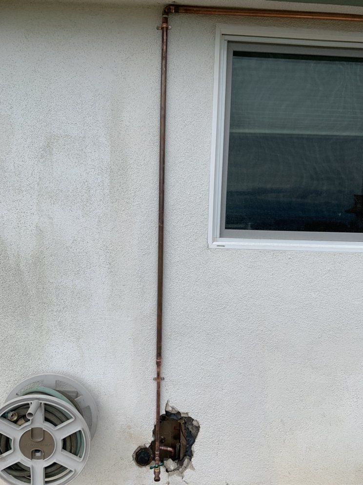 Side of a house with a wall-mounted utility meter, vertical pipe, window, and hose reel.