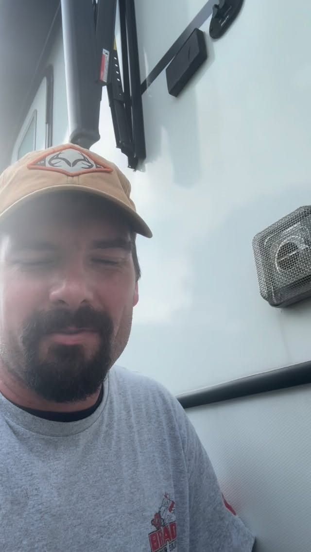 Man in tan cap and gray shirt smiling beside a white trailer with a mounted fan and hitch receiver.