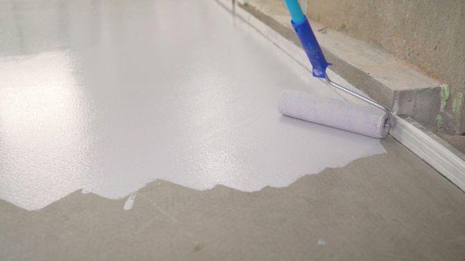 Paint roller applying white paint to a floor edge next to a wall inside a room.