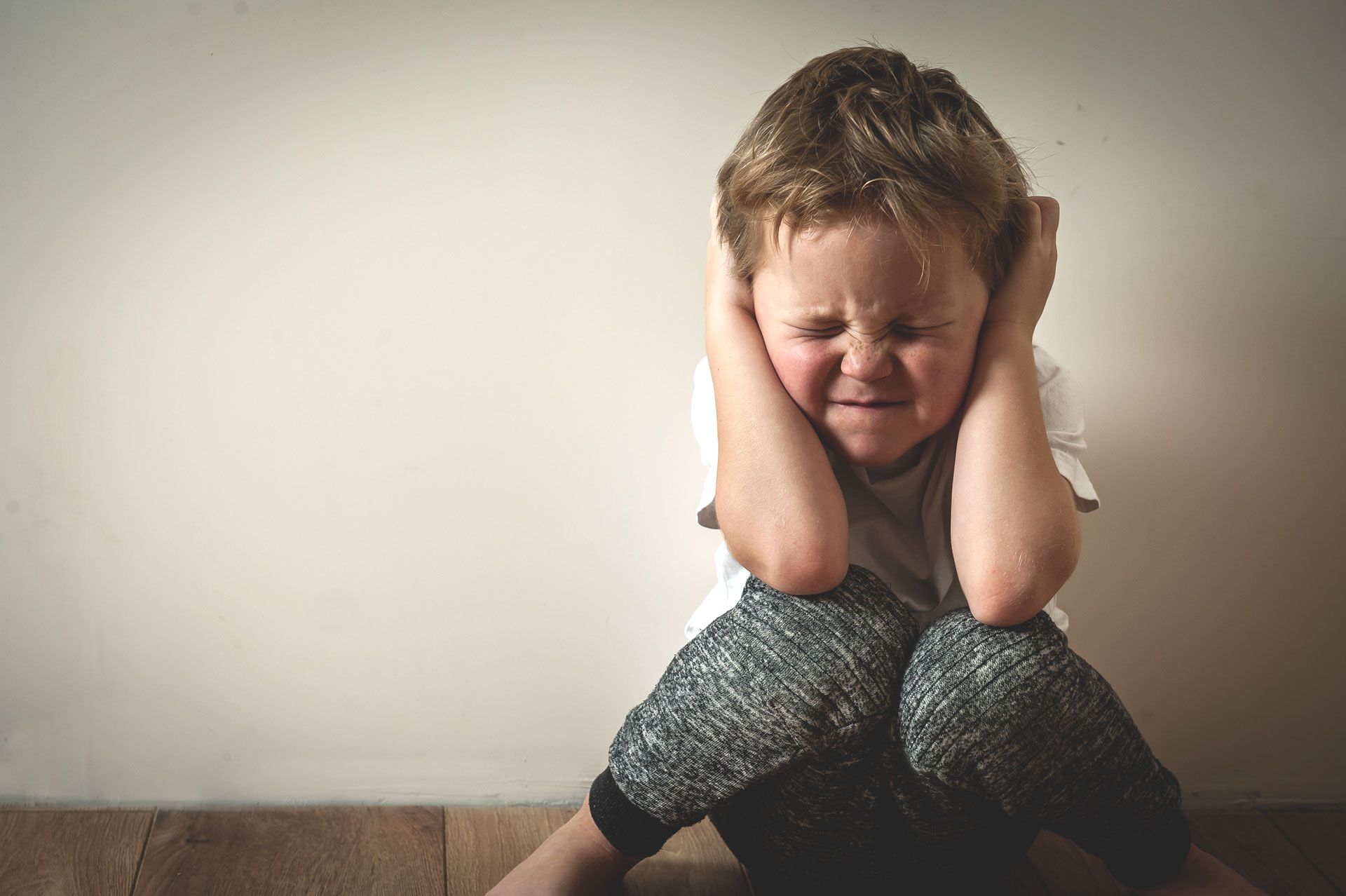 A young boy is sitting on the floor covering his ears with his hands.