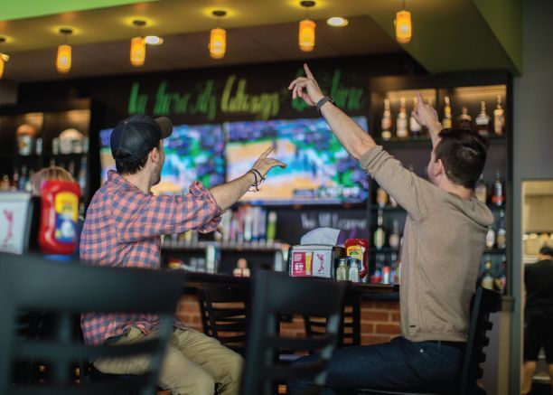 Two people in a bar raise their drinks while watching a TV screen at the counter.