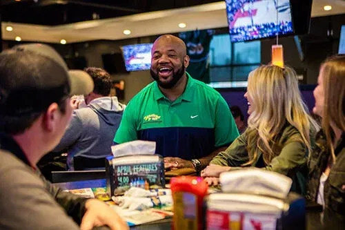Man smiling at a busy restaurant table with diners, menus, and condiment bottles in the foreground