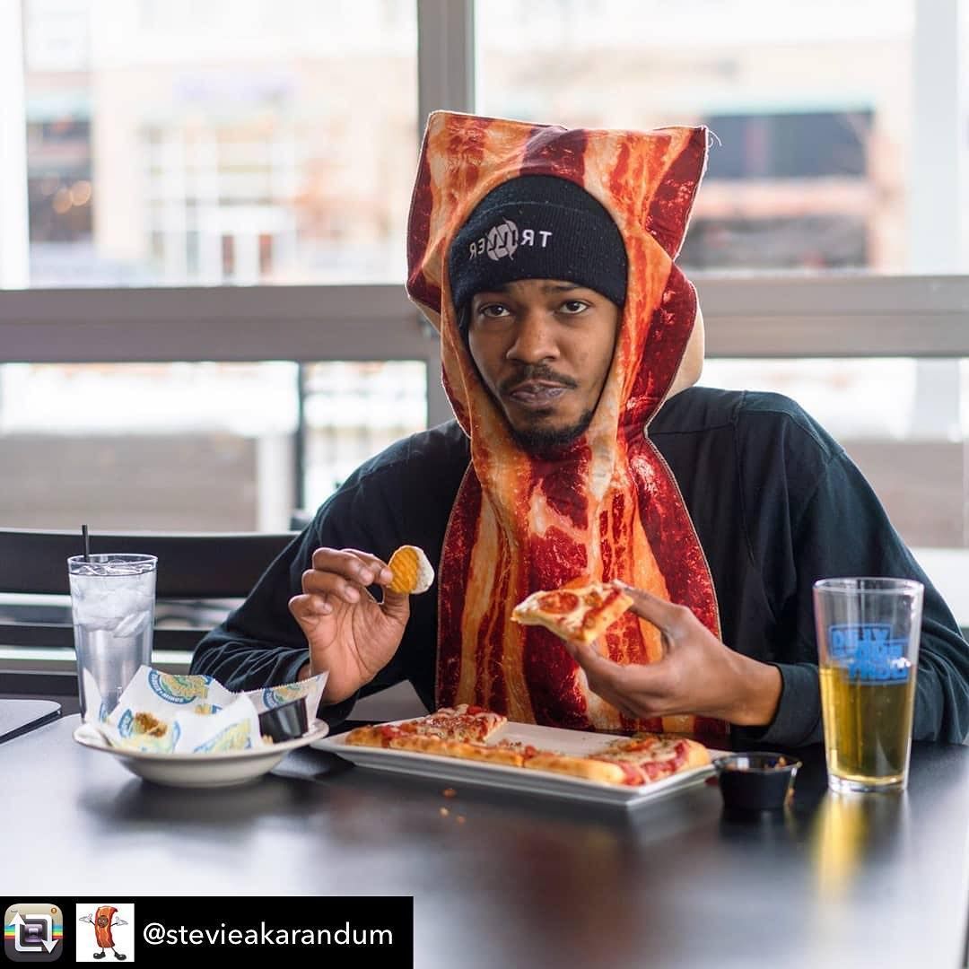 Person in a pizza-slice costume eating pizza at a table with drinks and a bowl, indoors by a window.