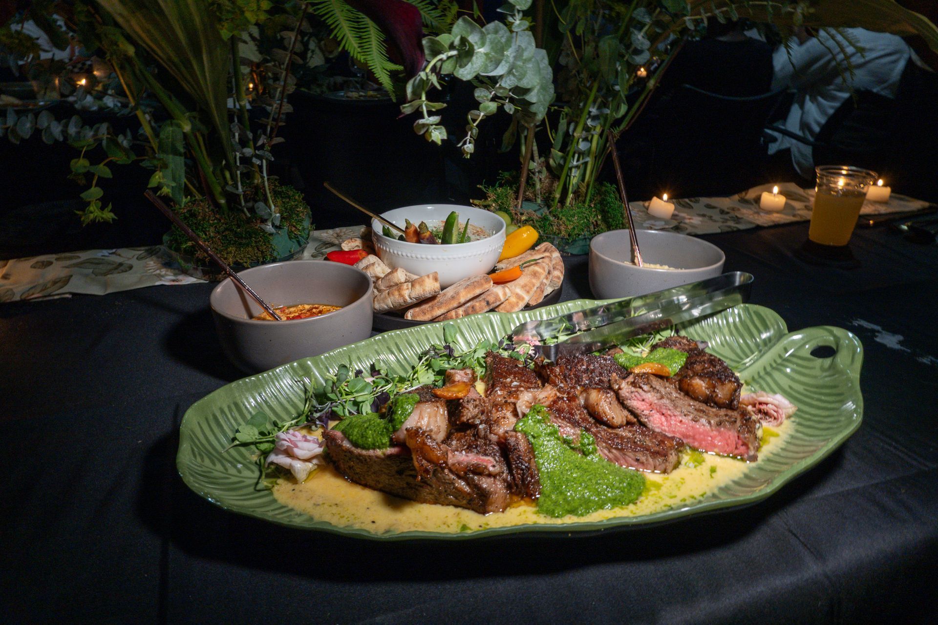 Plated grilled meat on a green leaf, with side bowls, on a candlelit black table setting.
