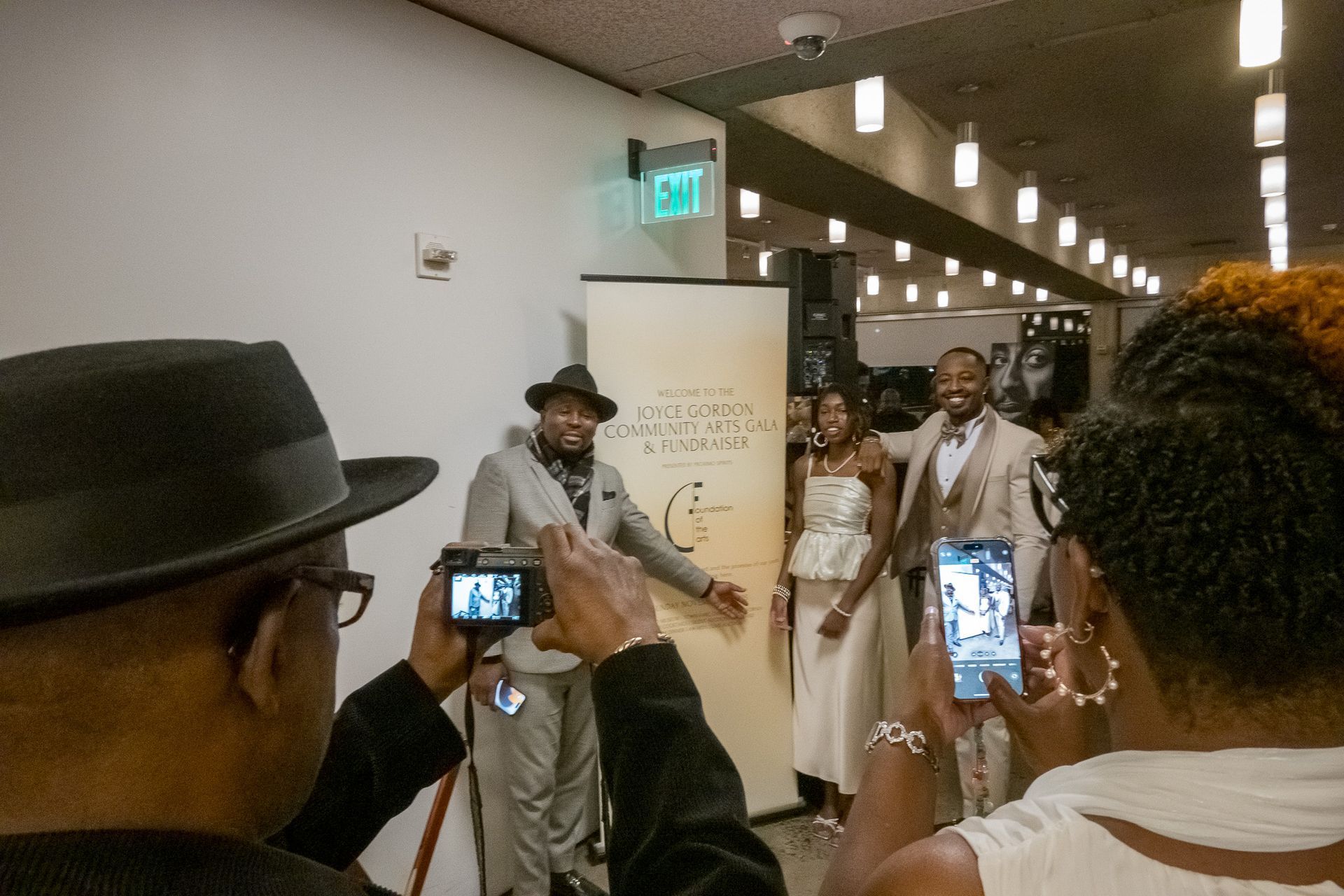 People filming performers in a hallway with bright ceiling lights and an EXIT sign