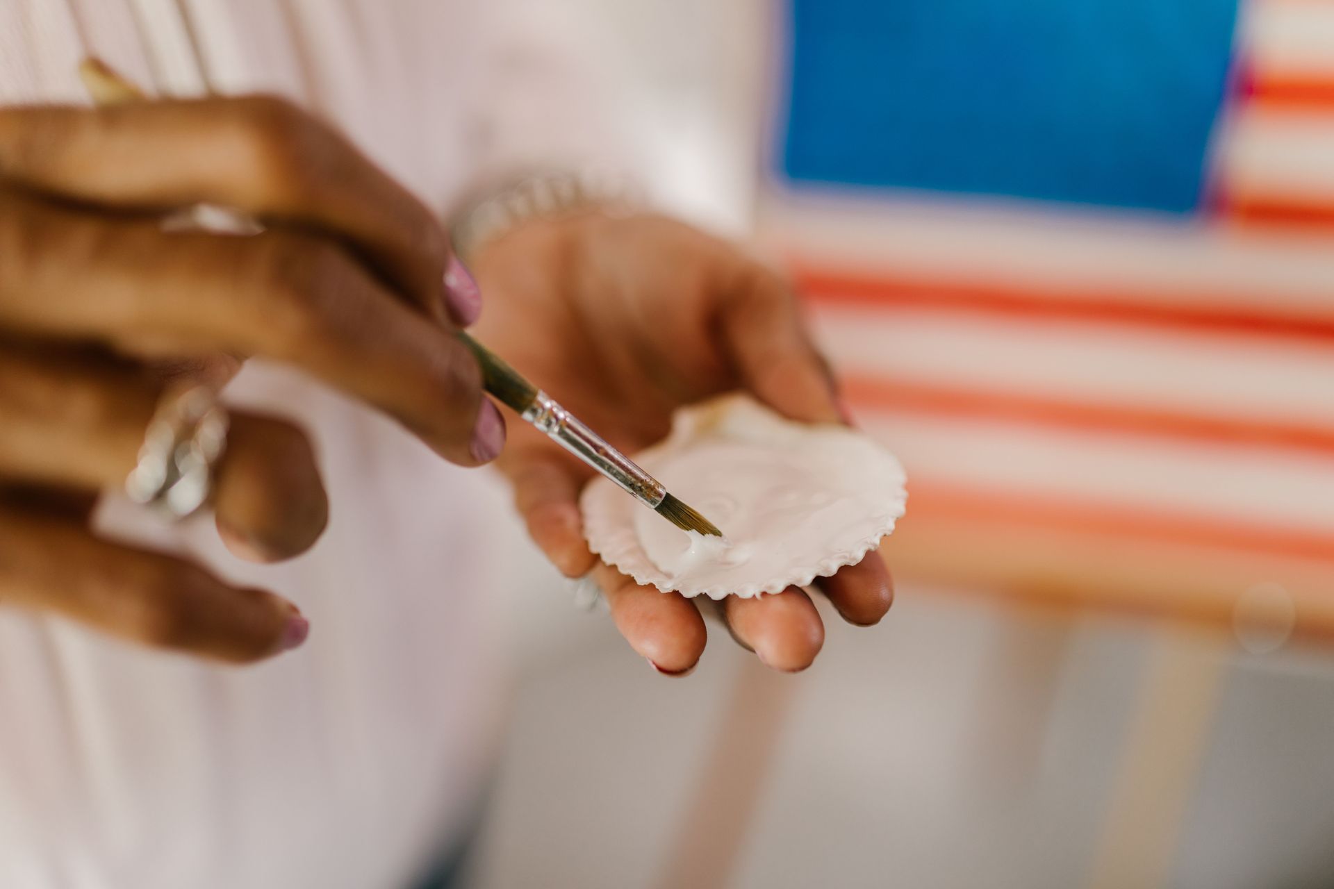 Hands holding a white shell with a paintbrush applying paint, with an American flag blurred in the background
