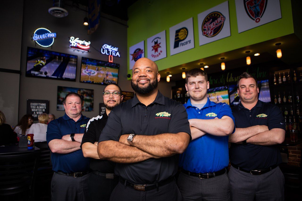 Five staff members stand with arms crossed in a sports bar with neon signs and framed jerseys.