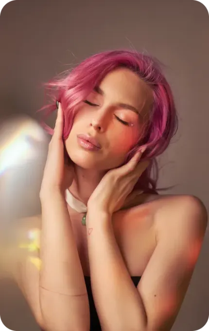 Woman with pink hair, eyes closed, hands on her cheeks. Soft lighting with color reflections.