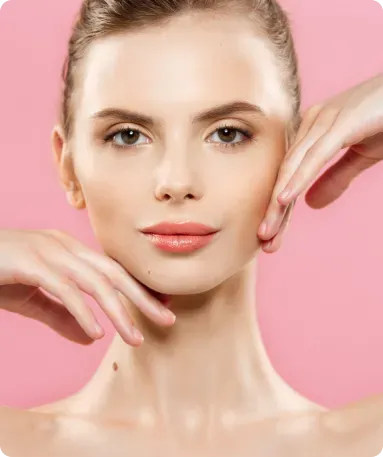 Woman with fair skin and brown hair, hands near face, posing in front of a pink background.