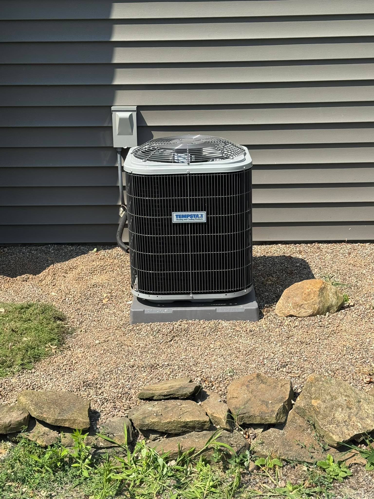 Central air conditioning unit against a gray house wall, surrounded by rocks and landscaping.