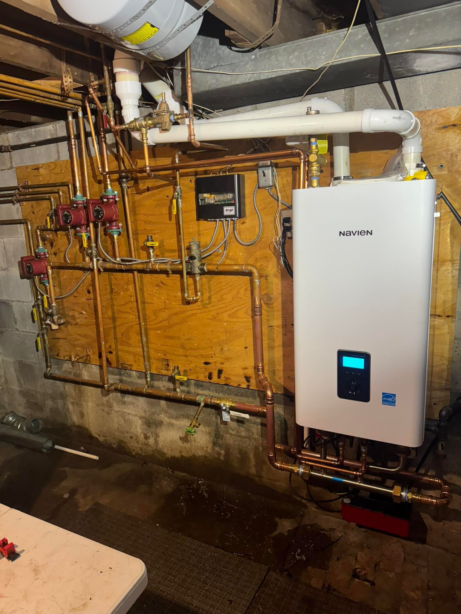 Copper pipes and a white water heater are mounted on a wood panel in a basement.