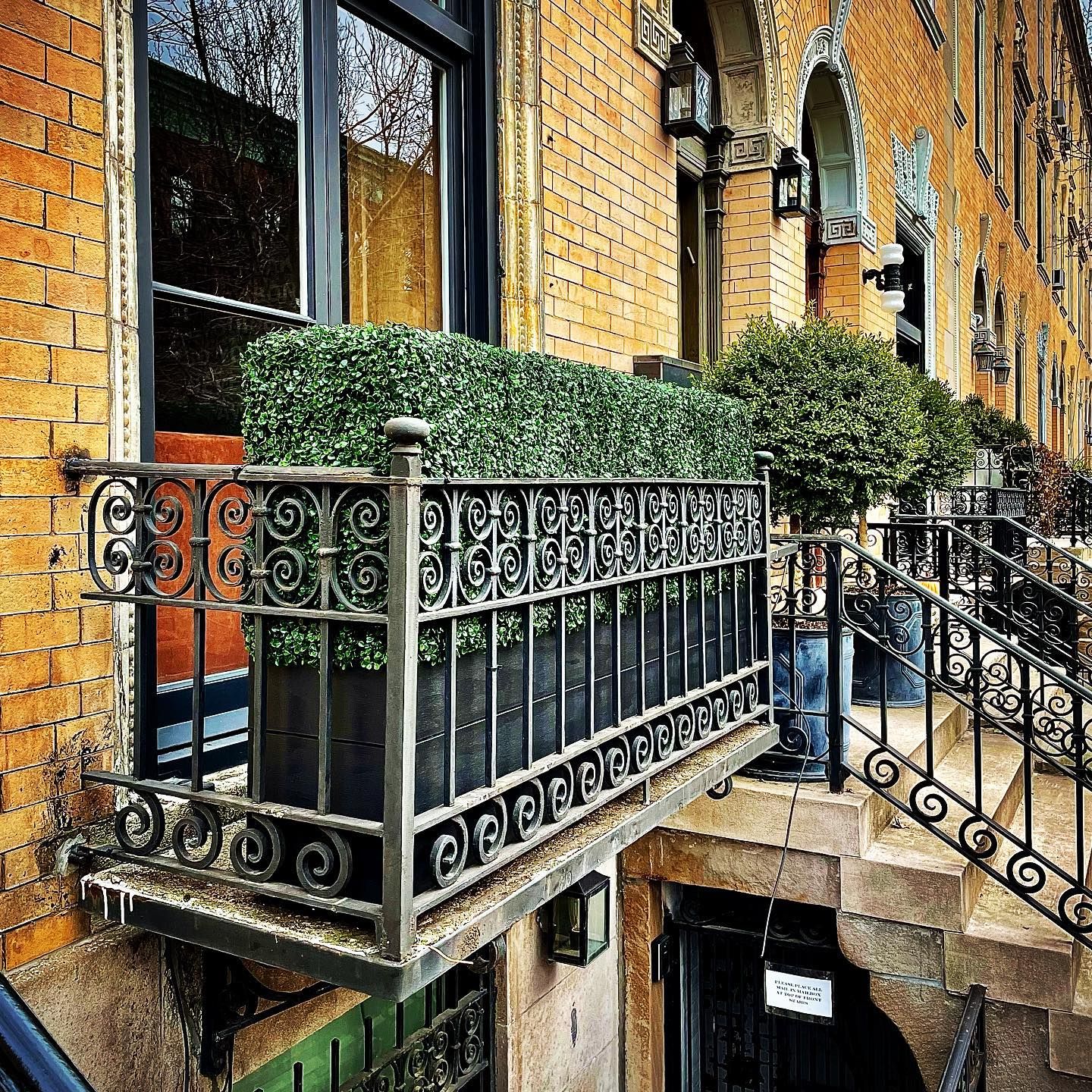 Wrought iron balcony with box hedge and decorative railing on brick building. Steps lead up to other balconies.