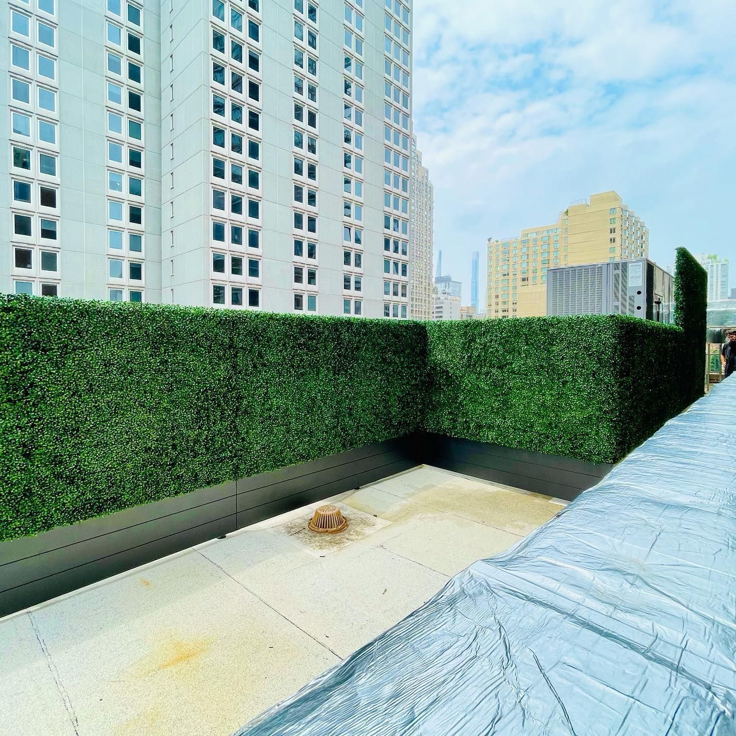 Rooftop with green hedge, concrete floor, buildings in background, cloudy sky.