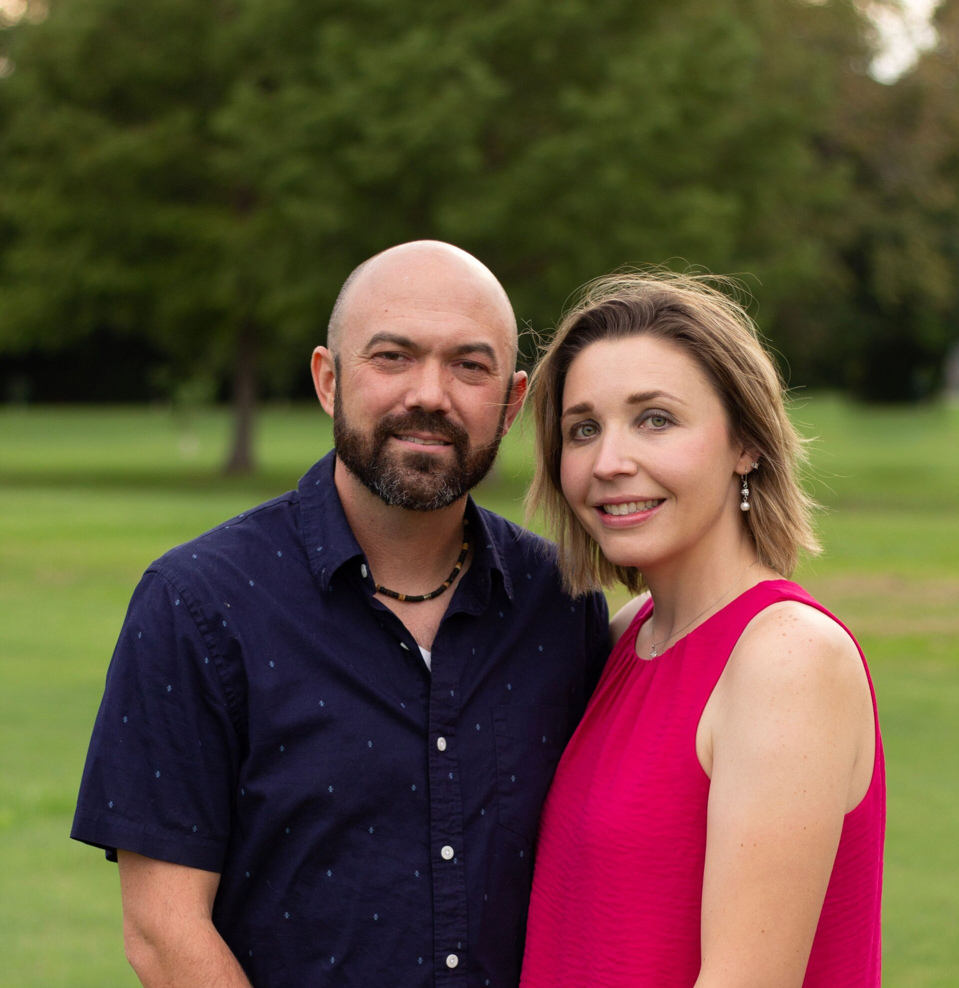 A man with a beard and a woman smile together outside. He wears a blue shirt; she wears a pink dress.