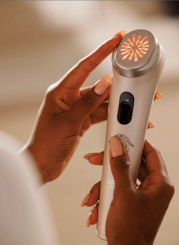 Person holding a facial treatment device with red light radiating from the head. Hands are visible.