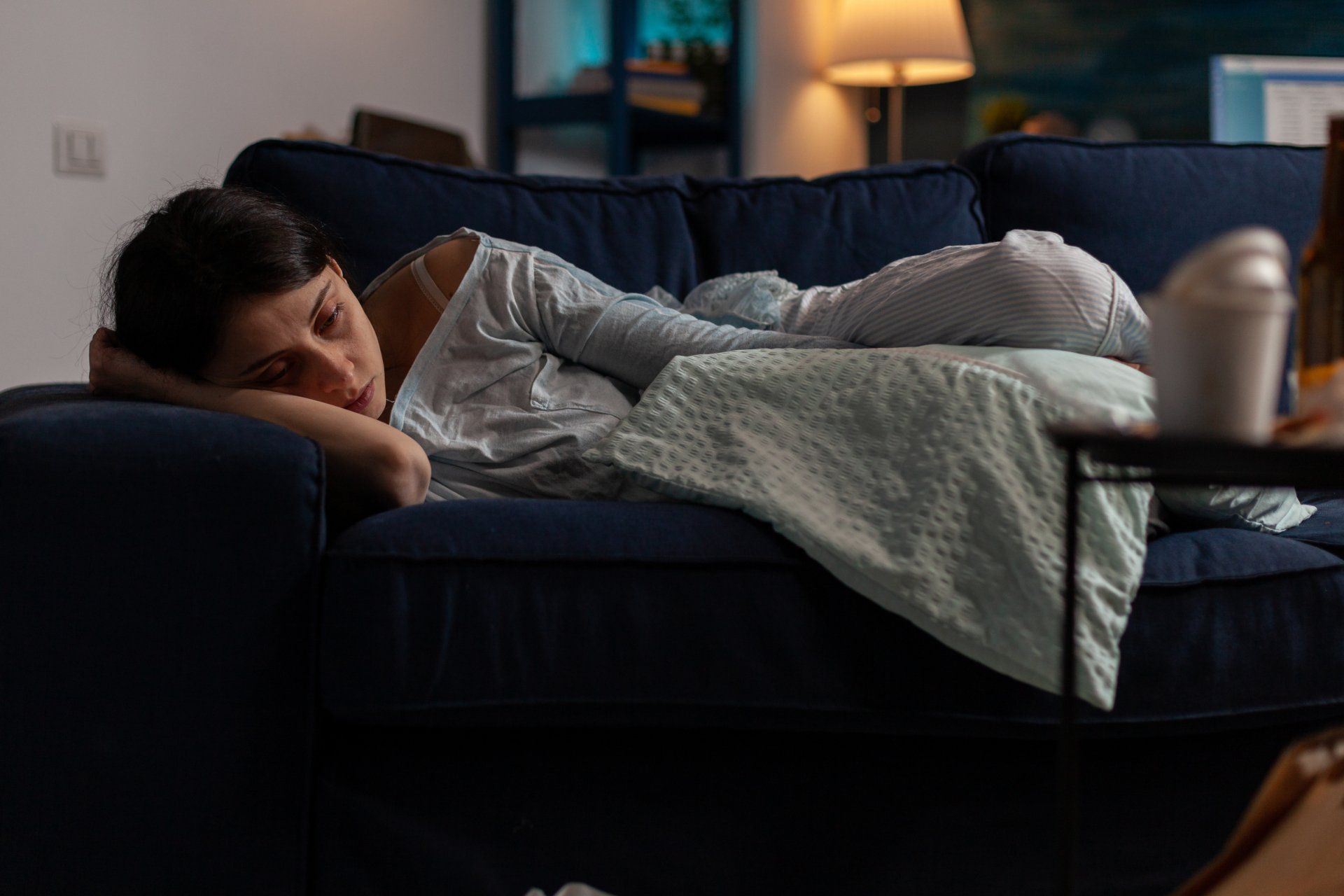 Woman asleep on a blue couch, arm draped over head. Soft blanket and lamp lit room.