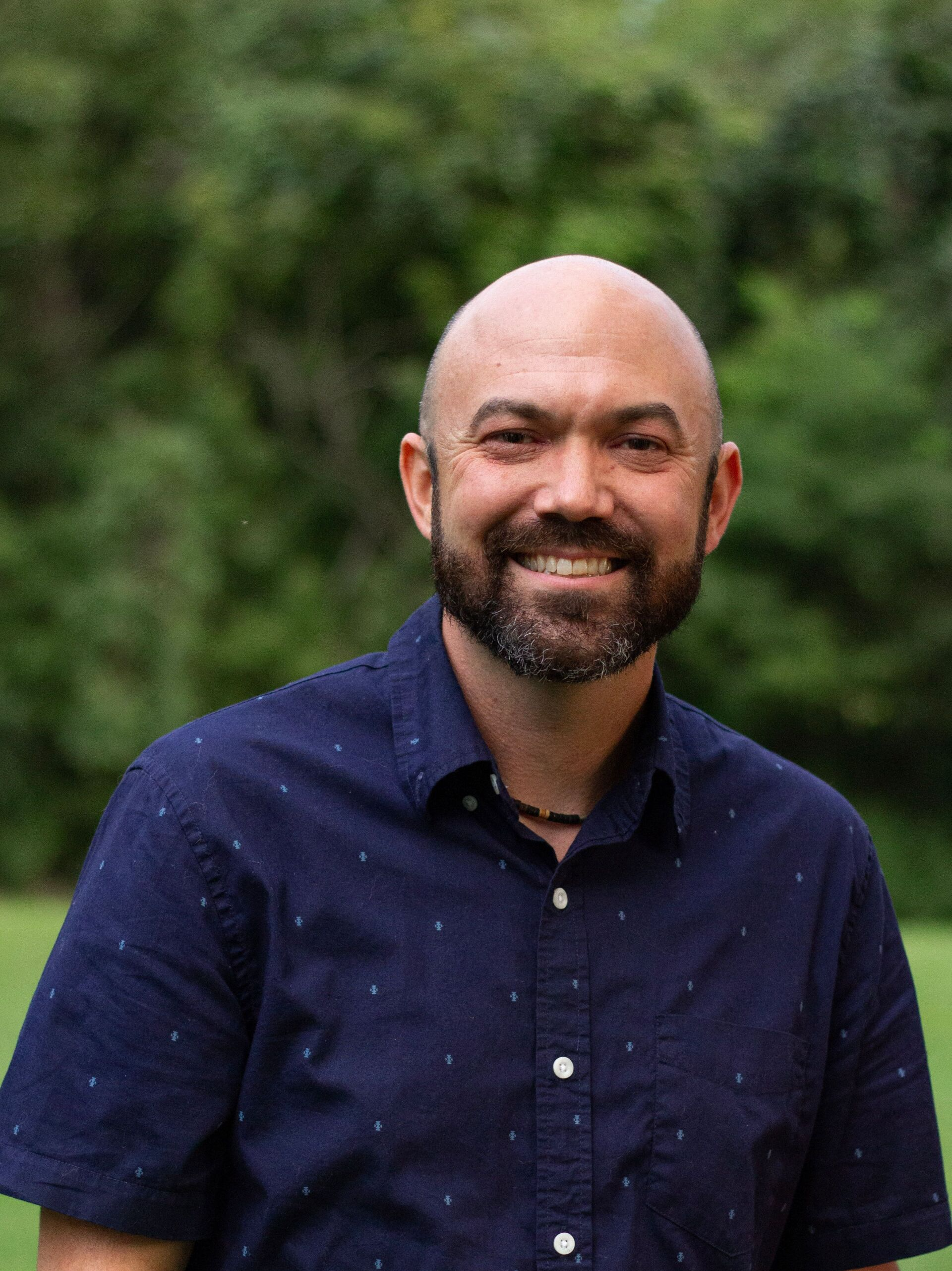 Smiling man with a bald head and beard, wearing a blue button-down shirt, in front of a green leafy background.
