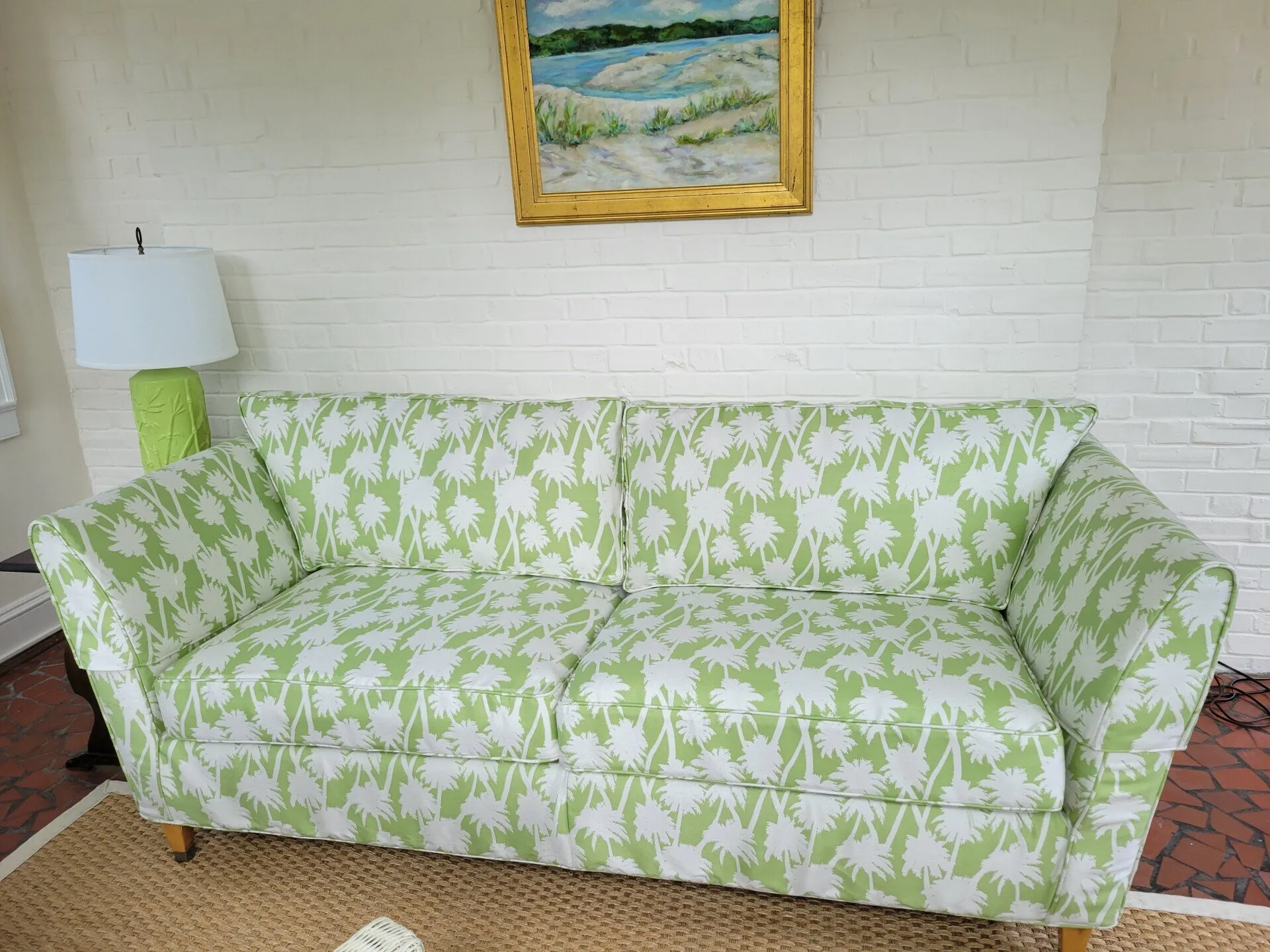 Green patterned couch against a white brick wall, with a landscape painting above and a green lamp beside it.