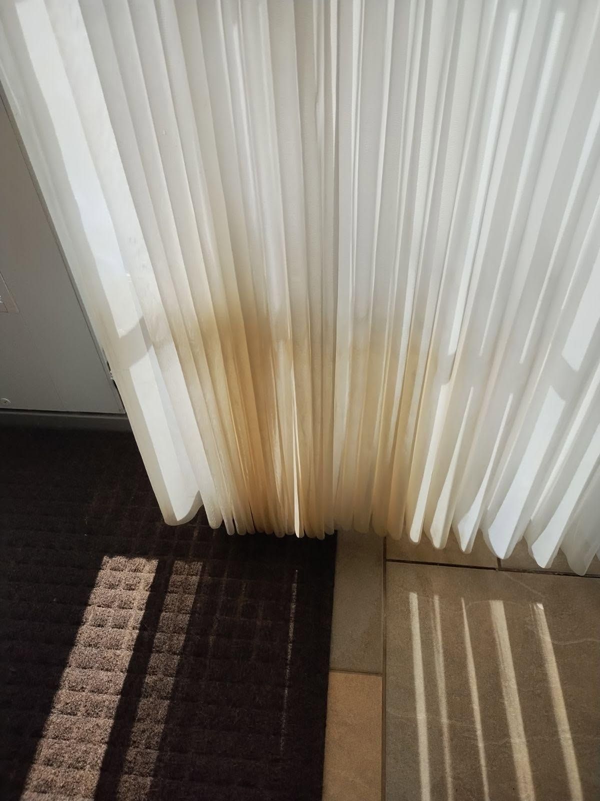 Sheer vertical blinds with a brown stain near the bottom beside a dark rug and sunlit floor.