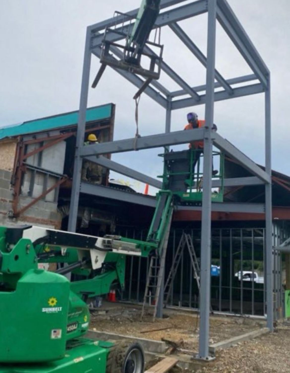 Workers on a green lift assembling a steel building frame at a construction site